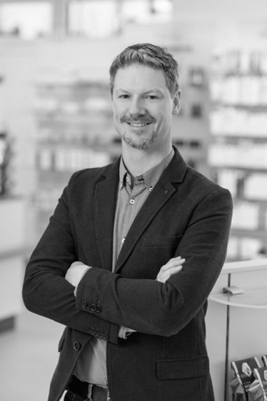Porträt eines freundlich lächelnden Mannes - Stefan Gritsch Inhaber der Apotheke Wildschönau - in Geschäftskleidung mit verschränkten Armen in der Apotheke.