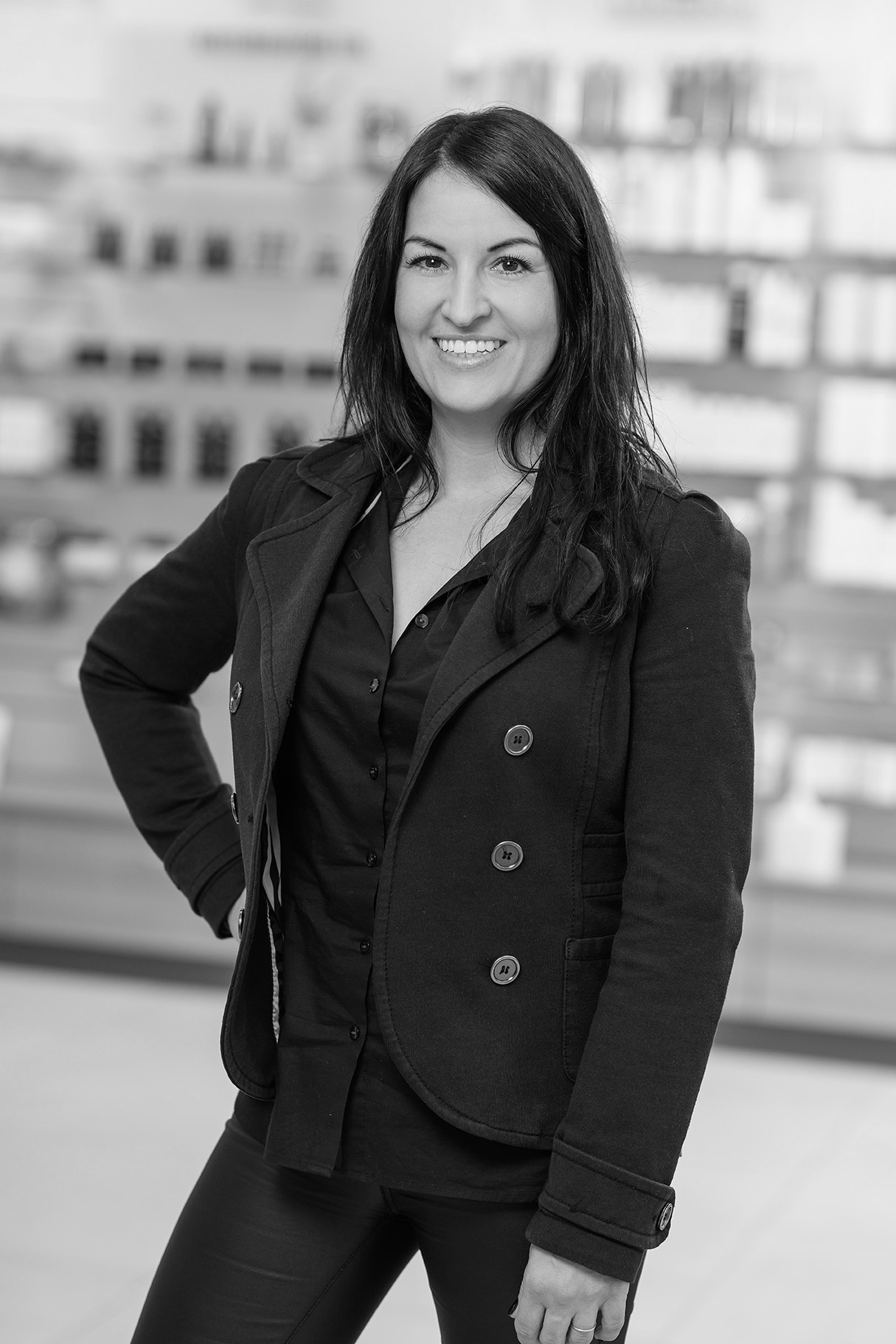 Mitarbeiterin Tamara Graustufen Porträt einer freundlich lächelnden Frau mit dunkler Kleidung, stehend vor einem Regal mit Produkten.